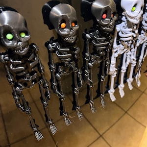 May include: Five small skeleton figurines with glowing orange and green eyes. The skeletons are black and white and are arranged in a row on a black surface.