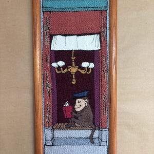 May include: A rectangular punch needle embroidery in a wooden frame depicting a monkey wearing a blue cap, sitting on a ledge and reading a red book. The scene features a dark purple interior, a yellow chandelier, and a white valance above.