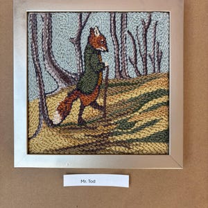 May include: Framed textile art depicting a fox in a green coat and walking stick, set in a forest scene. The artwork features a silver frame and the text "Mr. Tod" on a label below the image. The color palette includes shades of brown, green, and blue.