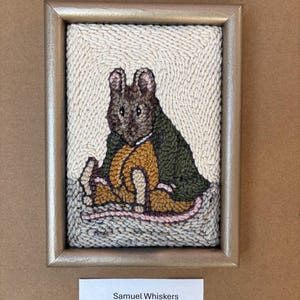 May include: Framed textile art featuring a seated brown rabbit wearing a green coat and yellow vest. The artwork is set against a cream-colored background and framed in a gold-toned frame. The text "Samuel Whiskers" is displayed below the artwork.