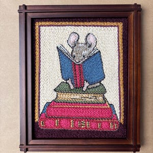 May include: A framed punch needle embroidery art piece featuring a gray mouse reading a blue book while sitting atop a stack of three books in shades of green, pink, and red. The artwork is enclosed in a dark brown wooden frame with a textured border.