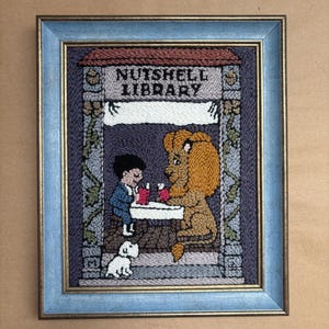 May include: A framed punch needle embroidery art piece titled NUTSHELL LIBRARY. The design features a person and a lion sitting at a table with a small white dog below. The embroidery uses purple, blue, yellow, and white yarn in a light blue frame.