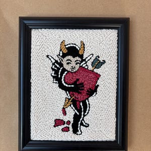 May include: Framed artwork featuring a devil-like figure with horns, wings, and a heart pierced by an arrow. The artwork is in a black frame, with a textured white background. The figure is in black, red, and gold.