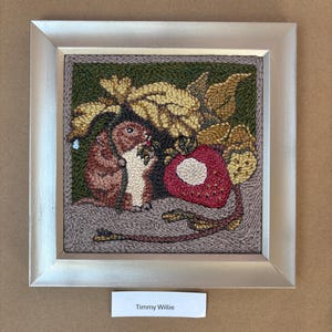 May include: A framed textile art piece featuring a detailed depiction of a rodent eating a berry next to a large red fruit. The artwork is framed in a silver-colored frame and includes the text "Timmy Willie" on a small white label.