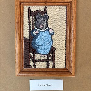 May include: Framed textile art featuring a pig wearing a blue dress, seated on a wooden chair. The artwork is in a wooden frame, with the text "Pigling Bland" on a white label below the frame.