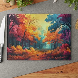 Puede incluir: Tabla de cortar rectangular de vidrio con una vibrante escena de bosque. La obra de arte representa un camino que serpentea a través de árboles con follaje colorido en tonos de rojo, naranja, amarillo y azul. La tabla está sobre una superficie gris con un cuchillo.