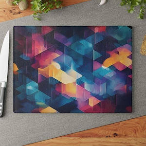 May include: Rectangular cutting board with an abstract geometric design. The pattern features overlapping shapes in shades of blue, purple, yellow, and pink. The board is set on a gray surface with a knife visible on the left.