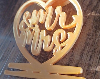 May include: A gold-coloured heart-shaped cake topper with the words "Mr & Mrs" in a decorative font. The topper is designed to hold a small cake or other dessert.