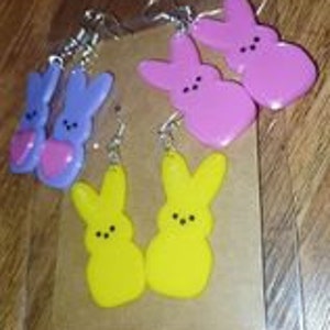 May include: Three pairs of dangle earrings featuring a Peeps bunny design. The earrings are made of plastic and come in yellow, pink, and purple.