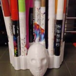 May include: White plastic desk organizer shaped like a hockey mask with a variety of colored markers and pens in the slots.
