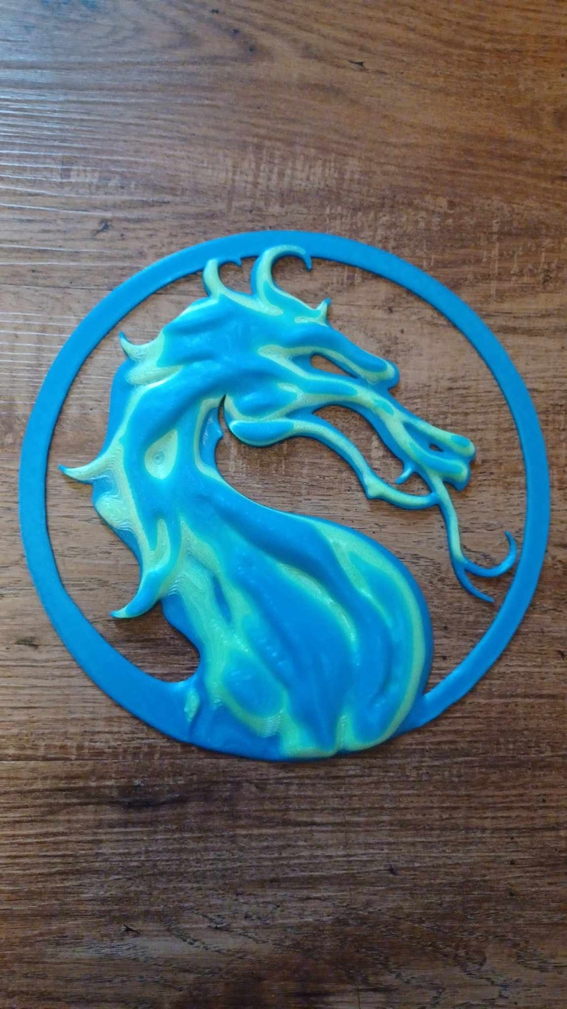 May include: A blue and green 3D printed dragon head in a circle. The dragon is facing right and has a light green and blue colour scheme.