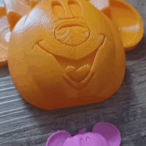 May include: Two 3D printed plastic molds of a cartoon mouse face. The larger mold is orange and the smaller mold is pink.