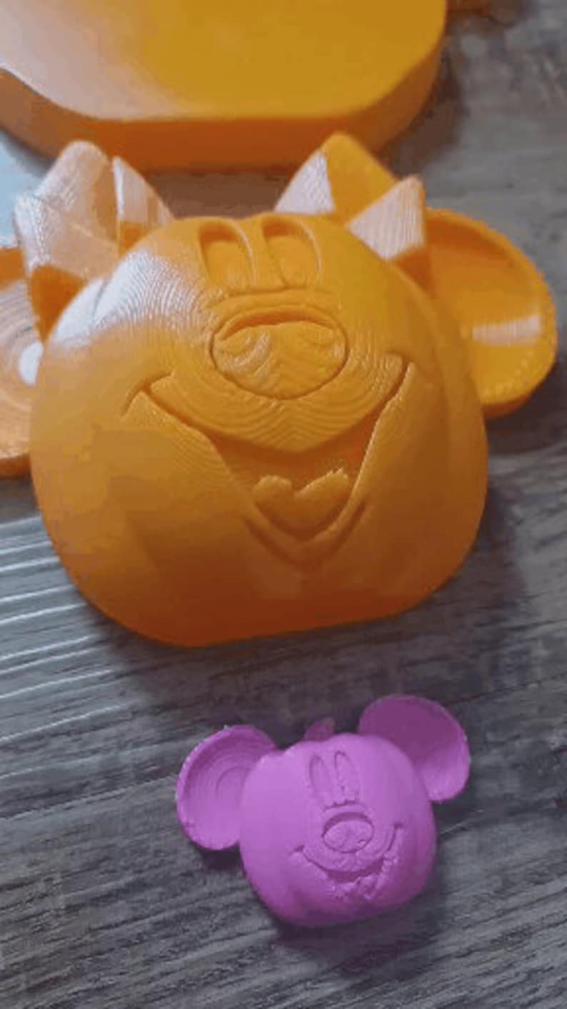May include: Two 3D printed plastic molds of a cartoon mouse face. The larger mold is orange and the smaller mold is pink.