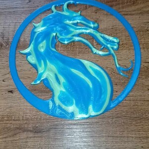 May include: A blue and green 3D printed dragon logo, resembling the Mortal Kombat logo, on a wooden background.