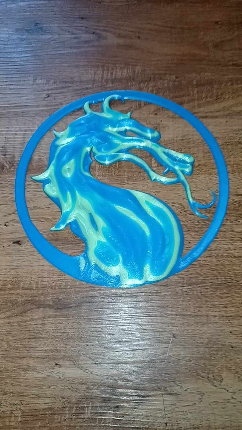 May include: A blue and green 3D printed dragon logo, resembling the Mortal Kombat logo, on a wooden background.