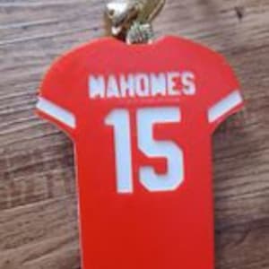 Puede incluir: Adorno de camiseta de fútbol roja y blanca con el nombre "Mahomes" y el número "15" en la espalda. El adorno está sujeto a una cuerda dorada.