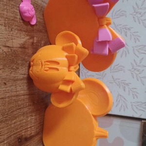 May include: Two orange 3D printed Mickey Mouse ears with pink bows. The ears are on a wooden surface.