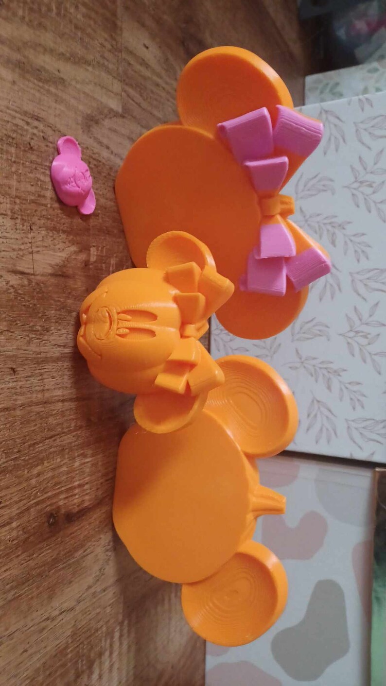 May include: Two orange 3D printed Mickey Mouse ears with pink bows. The ears are on a wooden surface.