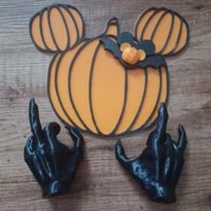 Könnte beinhalten: Eine Halloween-Dekoration mit einem großen orangefarbenen Kürbis mit Mickey-Mouse-Ohren und einer Fledermaus. Zwei schwarze Handskulpturen mit erhobenem Zeige- und kleinem Finger sind darunter positioniert.