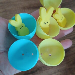 Pode incluir: Um Jogo de Caça aos Peeps com doces de marshmallow Peep amarelos dentro de ovos de Páscoa de plástico amarelos e azuis. O texto "PEEP HUNTING GAME" é exibido em letras verdes.