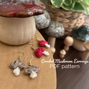 Könnte beinhalten: Gehäkelte Pilzohrringe in Rot und Braun mit weißen Akzenten. Die Ohrringe werden auf einer Holzoberfläche mit einer Pilzfigur im Hintergrund gezeigt. Der Text "Crochet Mushroom Earrings PDF pattern" wird unter den Ohrringen angezeigt.
