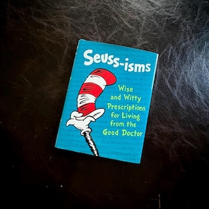 May include: A small, rectangular book titled "Seuss-isms" with a blue cover. The cover features the Cat in the Hat's red and white striped hat and the text "Wise and Witty Prescriptions for Living from the Good Doctor."