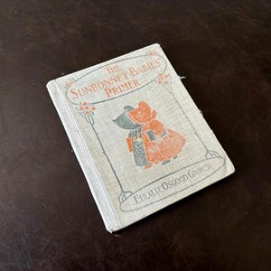 Pode incluir: Uma cópia vintage de "The Sunbonnet Babies Primer" de Eulalie Osgood Grover. O livro tem uma capa cinza clara com ilustrações de duas crianças com capotas e roupas laranja e verde. O livro está em uma superfície marrom escura.