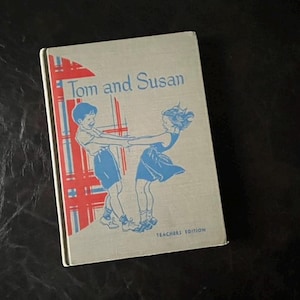 以下が含まれることがあります： 赤と白の格子柄の背景を持つビンテージな本の表紙。タイトル「トムとスーザン」は黒で印刷されています。青い服を着た男の子と女の子が手をつないで笑顔で描かれています。下部に「教師版」というテキストが黒で印刷されています。