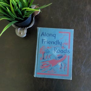 May include: A blue book with the title "Along Friendly Roads" on the cover. The cover features illustrations of people, cars, and trees in a whimsical style.
