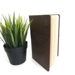 May include: A brown leather-bound book with cream-colored pages stands upright next to a small black potted plant with green grass-like leaves. The book's cover has embossed text. The background is white.