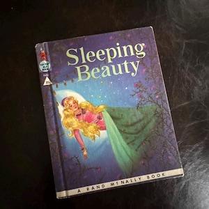 Puede incluir: Libro vintage "Sleeping Beauty" con una ilustración de portada de una princesa durmiente con cabello rubio largo, vestida con un vestido rosa y bajo una manta verde. El libro tiene una portada azul con el título en blanco y es un libro de Rand McNally.