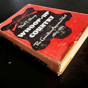 May include: A vintage book titled "WHOOP-UP COUNTRY" by Paul F. Sharp. The cover is red with black text and illustrations, including crossed rifles. The subtitle reads "The Canadian American West 1865-1885."