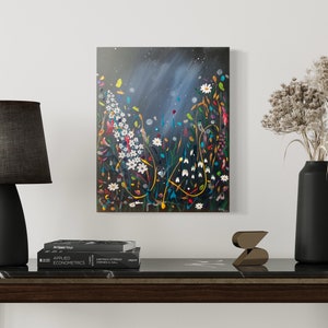 May include: An abstract floral painting with white daisies and colorful accents hangs above a dark wood table. A black lamp with a gray shade and a black vase with dried flowers are on the table.