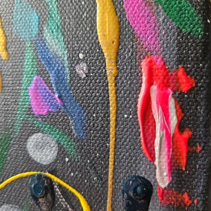 May include: Abstract art piece with a dark background and colorful paint strokes. The artwork features streaks of gold, red, pink, blue, and green paint, with small white and silver speckles throughout. The texture of the canvas is visible.