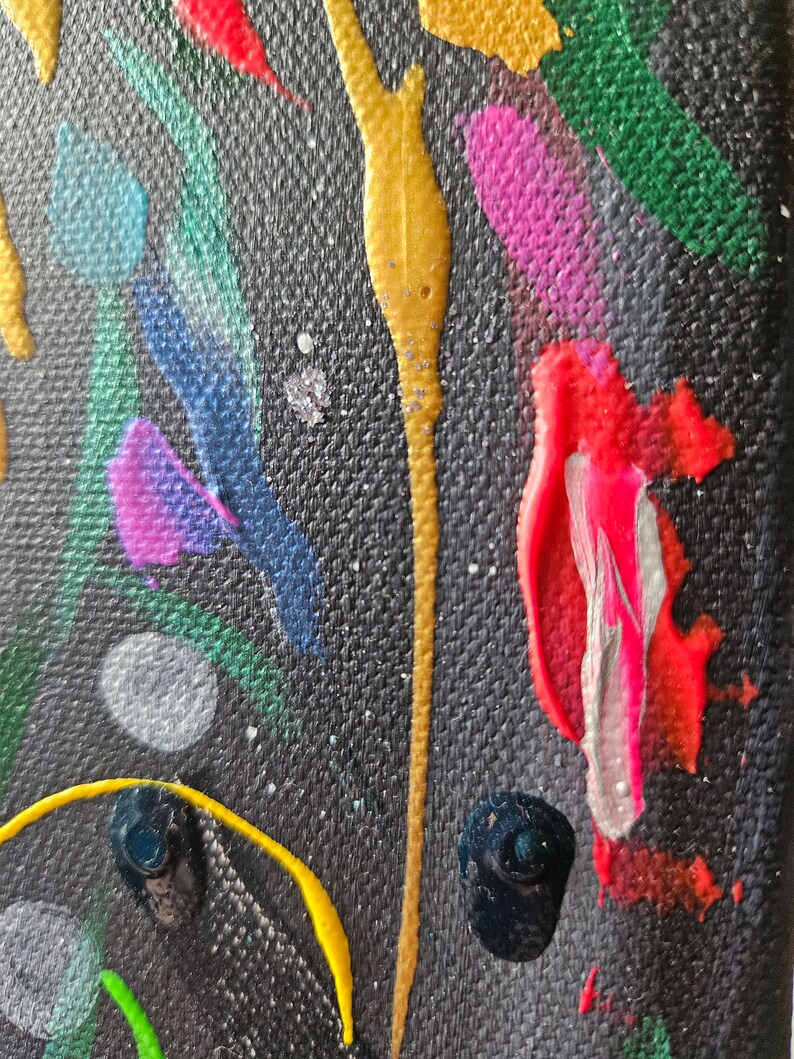 May include: Abstract art piece with a dark background and colorful paint strokes. The artwork features streaks of gold, red, pink, blue, and green paint, with small white and silver speckles throughout. The texture of the canvas is visible.