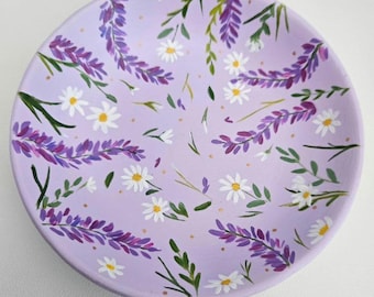 May include: A lavender and white floral design painted on a light purple ceramic dish. The dish is round and shallow, perfect for holding small items.