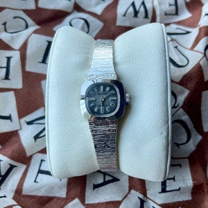 May include: A silver wristwatch with a square face and a textured band. The watch face is black with white markings and the text "Waltham 17 Jewels" printed on it.