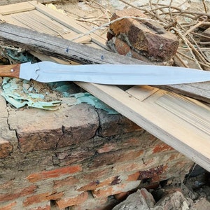 May include: A silver sword with a wooden handle lies on a piece of weathered wood. The wood is resting on a pile of bricks.