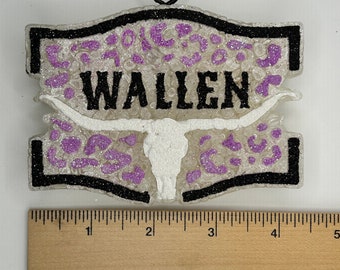May include: A white, glittery ornament with a black outline and a purple leopard print design. The ornament features a white bull skull and the word "WALLEN" in black lettering.