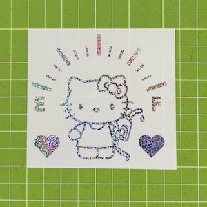 May include: A holographic vinyl decal featuring a Hello Kitty character holding a gas pump nozzle. The design includes a fuel gauge arc with the letters E and F, and two hearts on either side. The sticker has a reflective, multi-colored finish.
