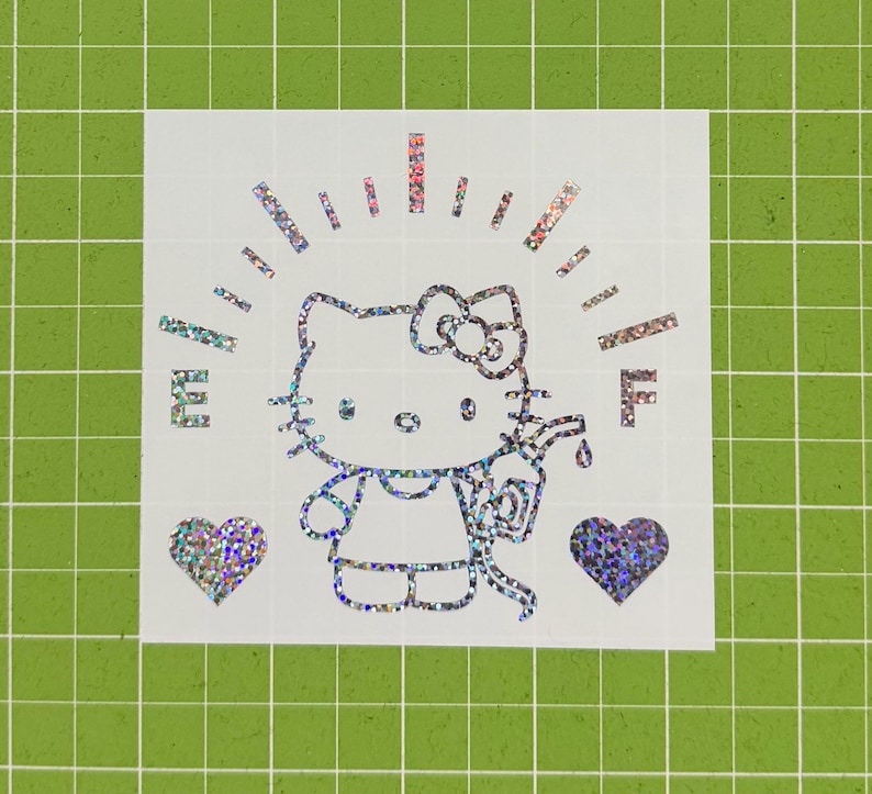May include: A holographic vinyl decal featuring a Hello Kitty character holding a gas pump nozzle. The design includes a fuel gauge arc with the letters E and F, and two hearts on either side. The sticker has a reflective, multi-colored finish.