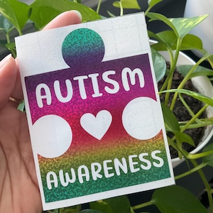 May include: A rectangular sticker featuring a puzzle piece shape with a holographic rainbow gradient background. The text AUTISM is printed in white at the top, with AWARENESS at the bottom, and a white heart icon centered between the two words.
