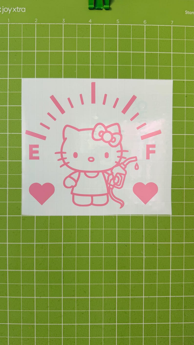 May include: A light pink vinyl decal featuring a cartoon cat character holding a gas pump nozzle. The design includes a fuel gauge arc with E and F markings, and two solid hearts on either side, all set against a white backing sheet.