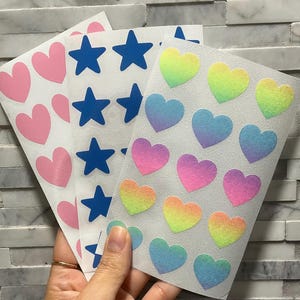 May include: Sheets of stickers featuring pink hearts, blue stars, and rainbow-colored hearts. The stickers are on white paper and held by a hand. The background is a light gray and white brick wall.
