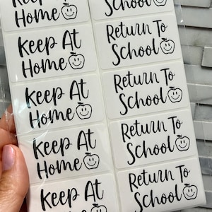 May include: A sheet of ten rectangular adhesive labels with black text and a small apple smiley face icon. Five labels read Keep At Home and five labels read Return To School in a cursive font. The stickers are arranged in two vertical columns.