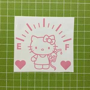 May include: A light pink vinyl decal featuring a cartoon cat character holding a fuel pump nozzle. The design includes a fuel gauge arc with the letters E and F, and two solid hearts on either side, all set against a white backing sheet.