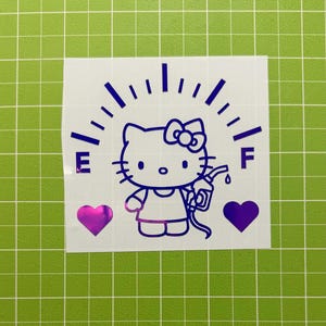 May include: A purple vinyl decal featuring a Hello Kitty character holding a gas pump nozzle, positioned below a fuel gauge arc with the letters E and F on either side, flanked by two solid purple hearts on a white backing sheet.