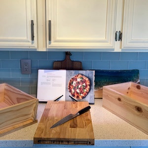 May include: Two wooden boxes with lids, a wooden chopping board, and a chef's knife on a kitchen counter. A cookbook with a pizza recipe is open on the counter.