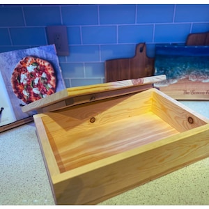 May include: A wooden box with a hinged lid, open to reveal an empty interior. The box is made of light-colored wood with a natural finish.