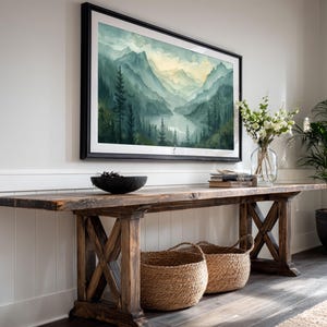 May include: A framed watercolor landscape painting depicting a mountain range with a river. The artwork is displayed above a rustic wooden console table with two woven baskets. A vase of white flowers and a potted plant are also present.